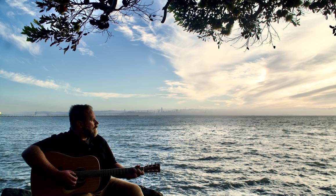 Robert Sullivan playing acoustic guitar beside the water